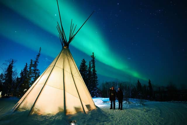 Two people standing beside a lit tipi beneath the northern lights